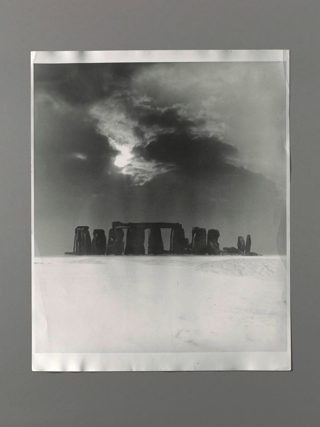 Black and white photograph. The dark posts and lintels of Stonehenge mark the transition from a stark, snowy field to a bright sunburst piercing dark clouds above.