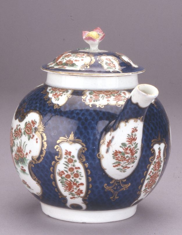 Teapot. The spout of a round-bodied ceramic teapot comes towards us, angled to our right, in this view. The body and lid are painted with brick-red flowers and butterflies in white cartouches, surrounded by gold edging against a navy-blue background. A pink-tipped flower sprouts from the lid.