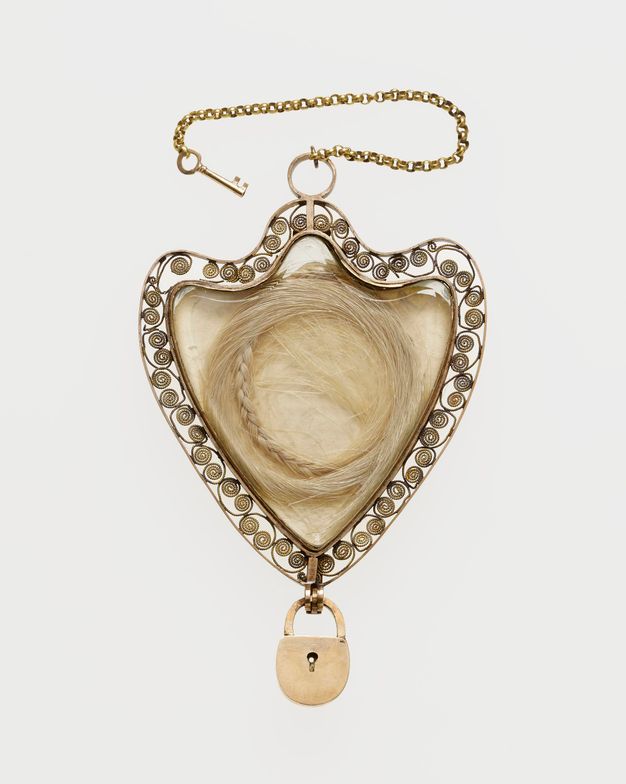 Jewellery. A shield-shaped pendant contains a braided locket of pale hair. The edge is surrounded by scrolling filigree. A miniature padlock hangs from the bottom and a tiny skeleton key is attached by a chain to the top.