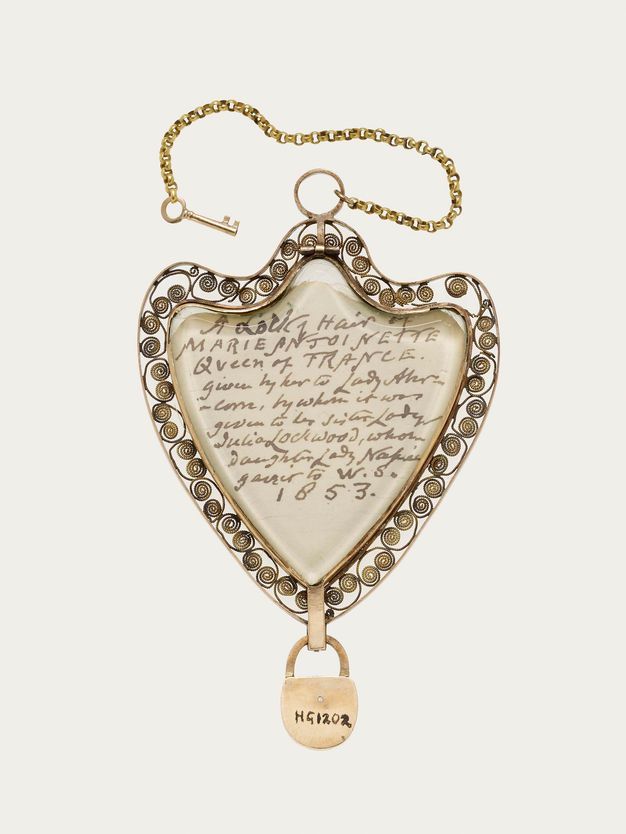 Jewellery. A shield-shaped pendant is surrounded by scrolling filigree. Writing within begins, “A Lock of Hair of Marie Antoinette Queen of France” and is dated 1853. A miniature padlock hangs from the bottom and a tiny skeleton key is attached by a chain to the top.